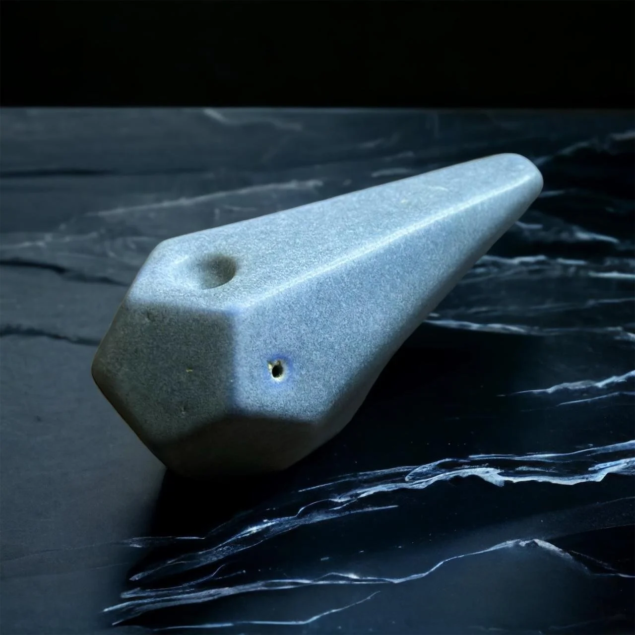 Pipa de cerámica gris con forma geométrica y acabado mate, colocada sobre una superficie oscura con vetas que recuerdan a la piedra o el mármol.