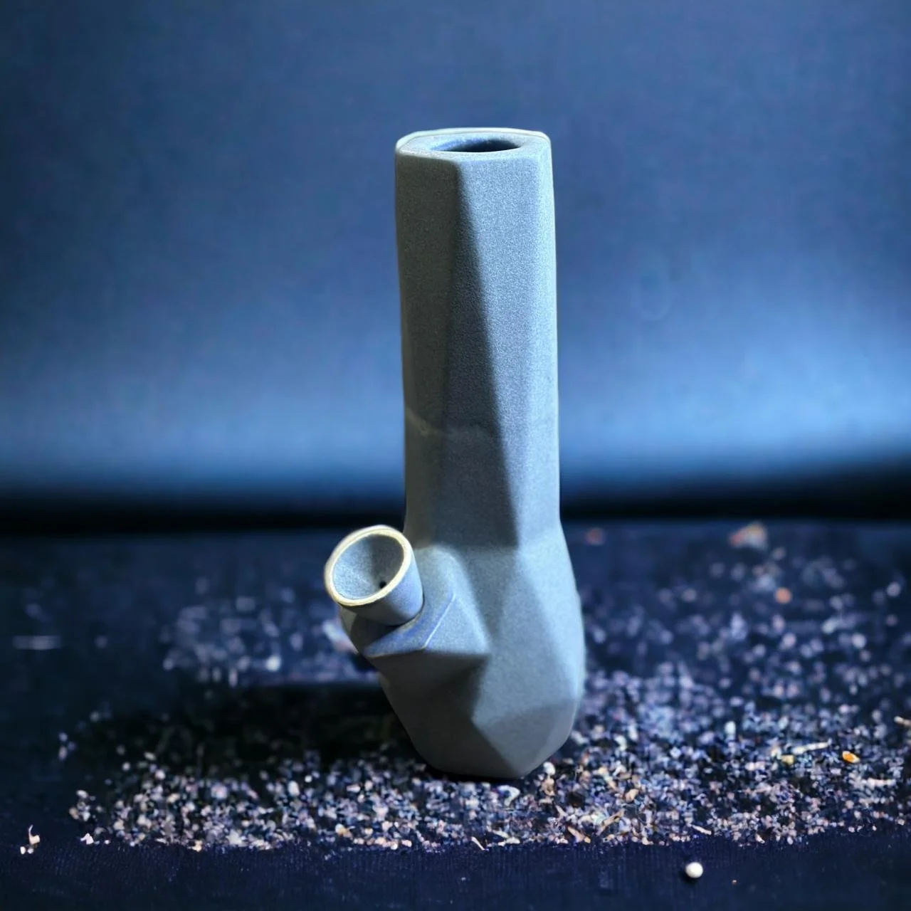Bong de cerámica gris con diseño geométrico y acabado mate, colocado en posición vertical sobre una superficie oscura con pequeñas partículas, con fondo azul desenfocado.