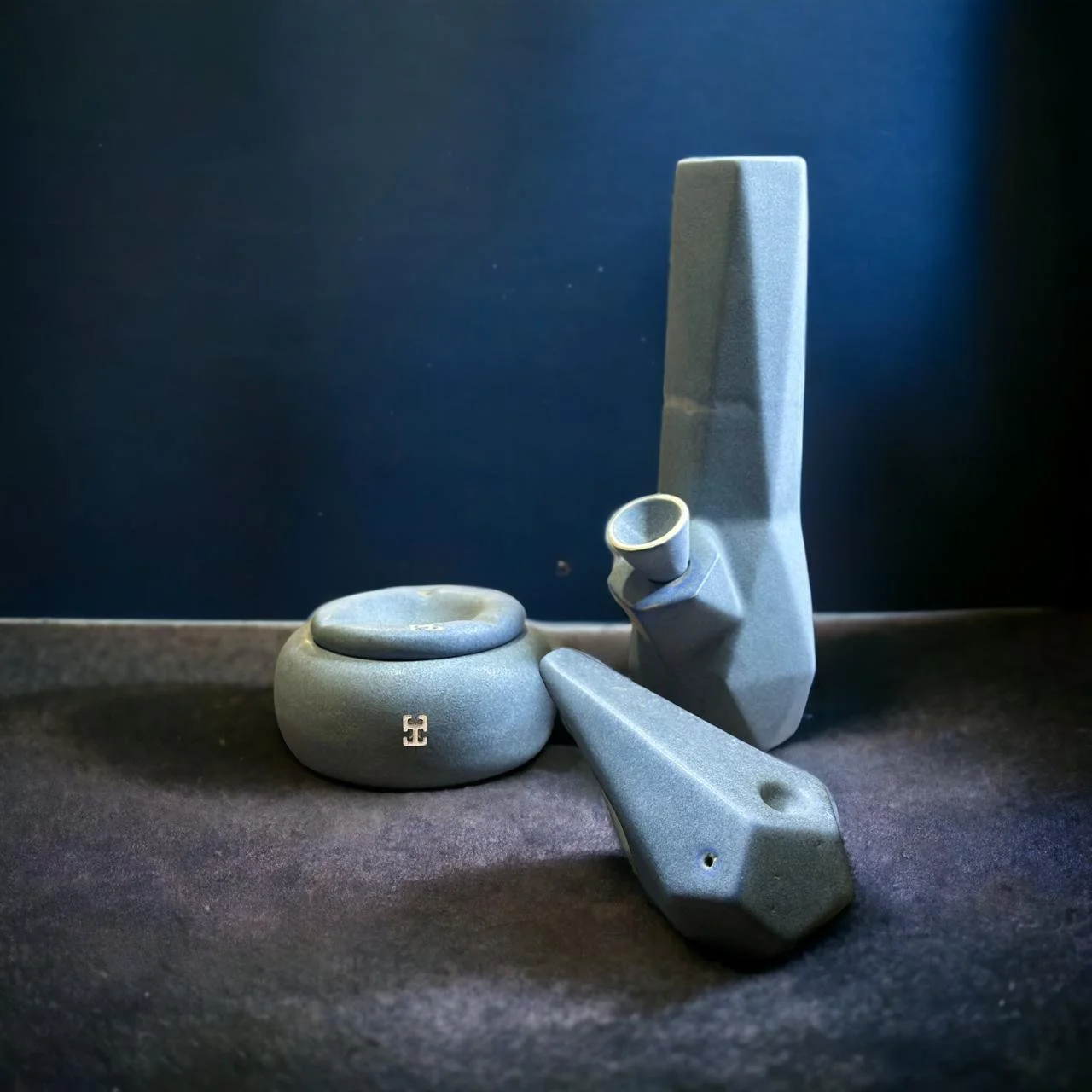 Conjunto de objetos de cerámica gris con diseño geométrico: un bong alto, una pipa y un recipiente redondo con tapa, colocados sobre una superficie de piedra en un interior