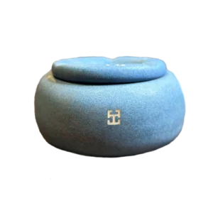 Caja de cerámica artesanal de forma redondeada en tono azul mate, con tapa y un pequeño símbolo decorativo en el frente.