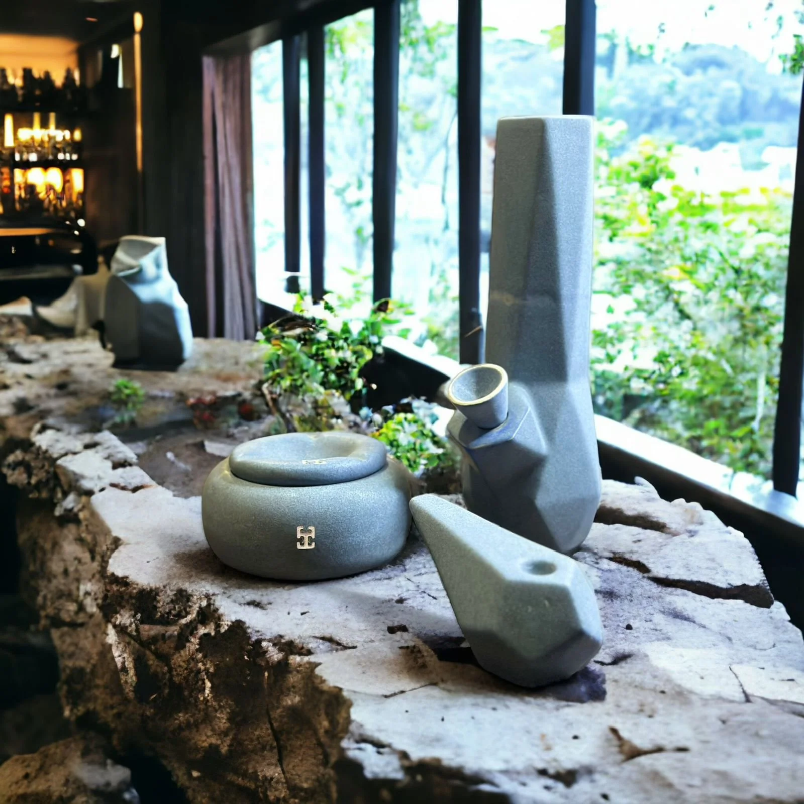Conjunto de objetos de cerámica gris con diseño geométrico: un bong alto, una pipa y un recipiente redondo con tapa, colocados sobre una superficie de piedra en un interior iluminado por grandes ventanales con vista a vegetación.