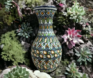 Jarrón de cerámica artesanal decorado con patrones geométricos en tonos verde, amarillo y azul, colocado entre plantas suculentas y musgo.