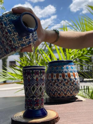 Mano vertiendo una bebida desde una jarra de cerámica artesanal en una taza decorada, ambas con patrones geométricos en tonos azul y multicolor, en un entorno exterior con plantas y cielo azul.